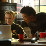 Medium shot of Kristen Bell as Veronica sitting at desk using laptop computer as Percy Daggs III as Wallace looks over her shoulder.