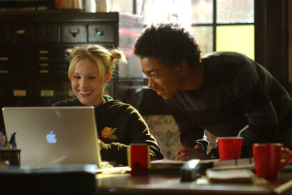 Medium shot of Kristen Bell as Veronica sitting at desk using laptop computer as Percy Daggs III as Wallace looks over her shoulder.