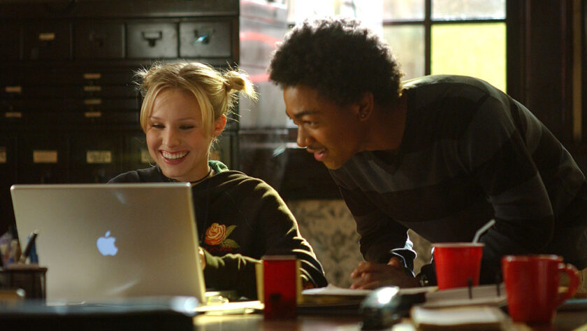 Medium shot of Kristen Bell as Veronica sitting at desk using laptop computer as Percy Daggs III as Wallace looks over her shoulder.